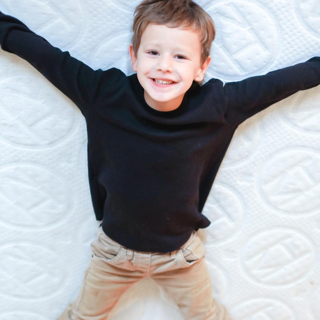 Smiling little boy wearing a black sweater and beige pants doing a snow angel position across a 6" Haven Jr mattress